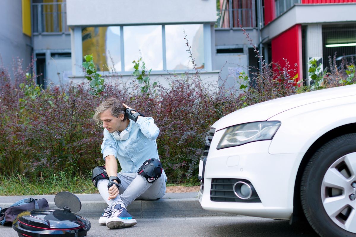 Car accident concept. Young student male man sitting on road with knee and hand protection with eco electric unicycle and taken injury in car accident with white gasoline diesel car. Safe road concept